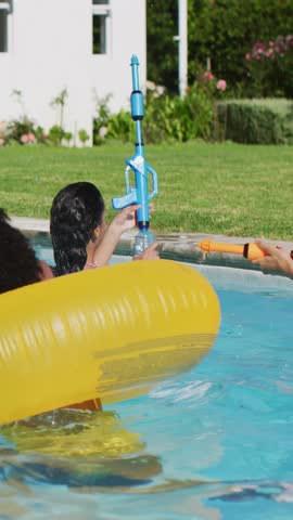 Vertical video: Offscreen orange blaster urging woman pumping blue blaster as icons rising for ads. Poolside, backyard, watergun, inflatable, sunny, vibrant, vertical video