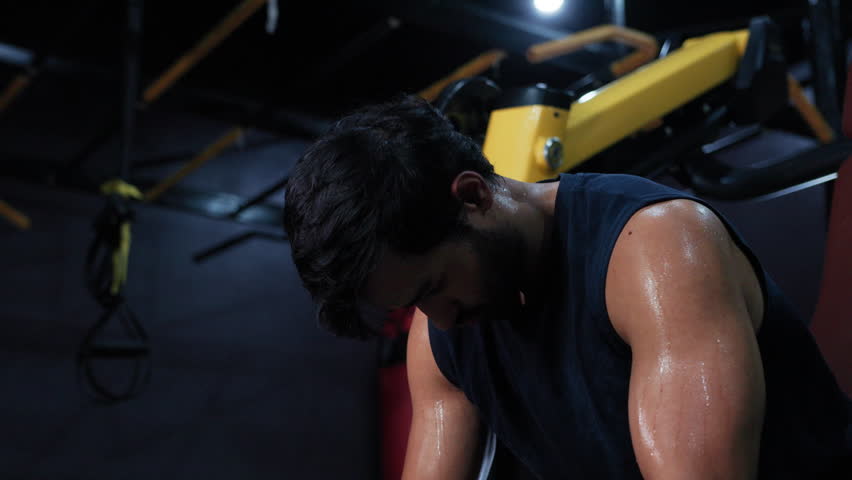 Tired Athlete Taking a Pause During Gym Workout Session, Man Resting on Gym Equipment Between Exercises, recovery, workout break, and athletic concentration.