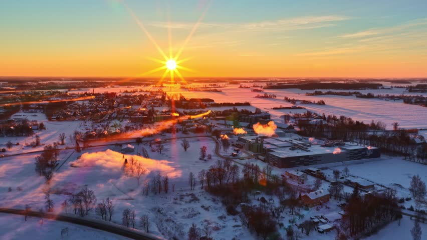 Aerial winter sunrise over a snowy town and frozen countryside, with golden sun rays, steam plumes, and glowing light across the cold landscape.