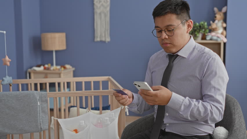 Man in shirt and tie holds smartphone and creditcard while seated beside a baby crib inside building; concerned parent responsibility.