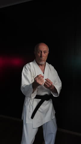 Against the background of flashing red and green lights, an old male athlete practices punches and kicks in motion