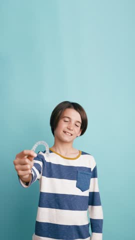 Teenager in a striped shirt holding a transparent orthodontic retainer. The young patient is smiling against a solid blue background. Vertical