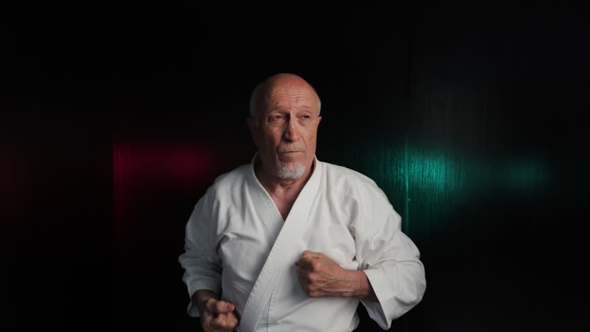 Against a background of flashing red and green lights, an old male athlete throws punches and blocks with his hands