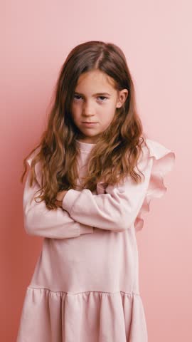 Unhappy young girl with arms crossed feeling offended and sulking. Displeased child expressing negative emotions on a pink background. Vertical