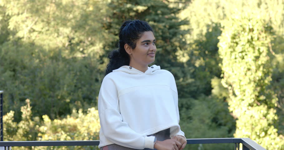 Adult female on balcony metal rail wearing hoodie reacting to camera tightening turning and smiling. Woman, terrace, balustrade, foliage, trees, sunlit, casual