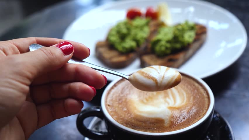 Woman with red manicure holding a small spoon with creamy latte foam over a cup of fresh cappuccino, with a plate of avocado toast visible in the blurred background of a cozy coffee shop