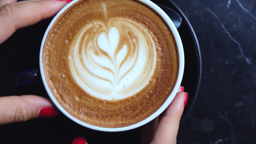 Female hands with red nail polish gently spinning a cup of hot cappuccino with beautiful flower shaped latte art on a black marble table, creating a cozy and relaxing cafe atmosphere