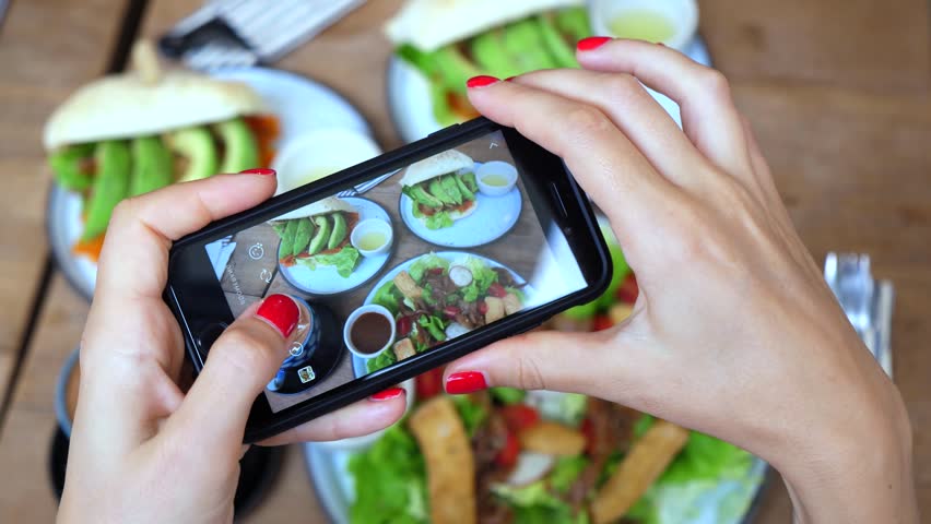 Close up view of female hands with red nail polish taking a photo of healthy vegan food with a mobile phone in a restaurant. Blogger lifestyle creating content for social media