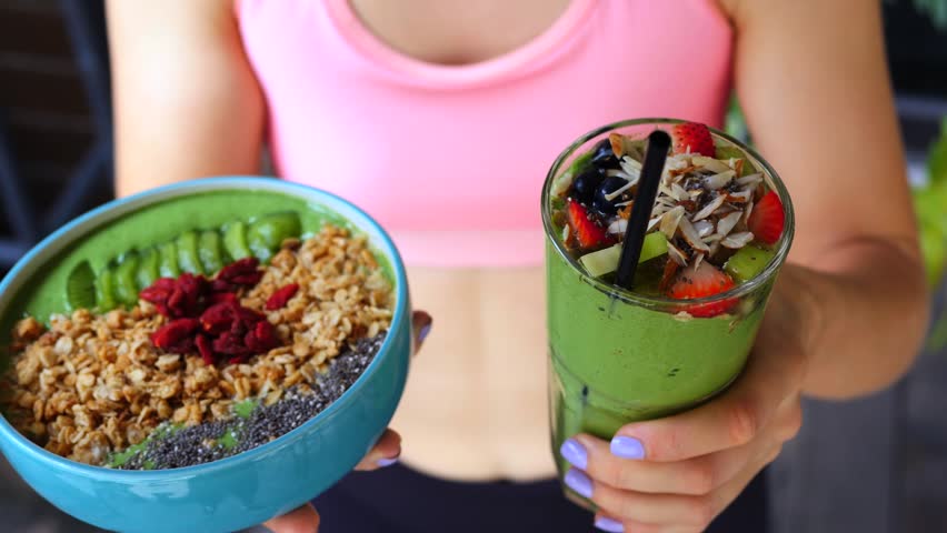 Woman in sportswear holding a healthy green breakfast smoothie bowl with granola, goji berries, and chia seeds, and a fresh spinach smoothie drink topped with fruits and nuts