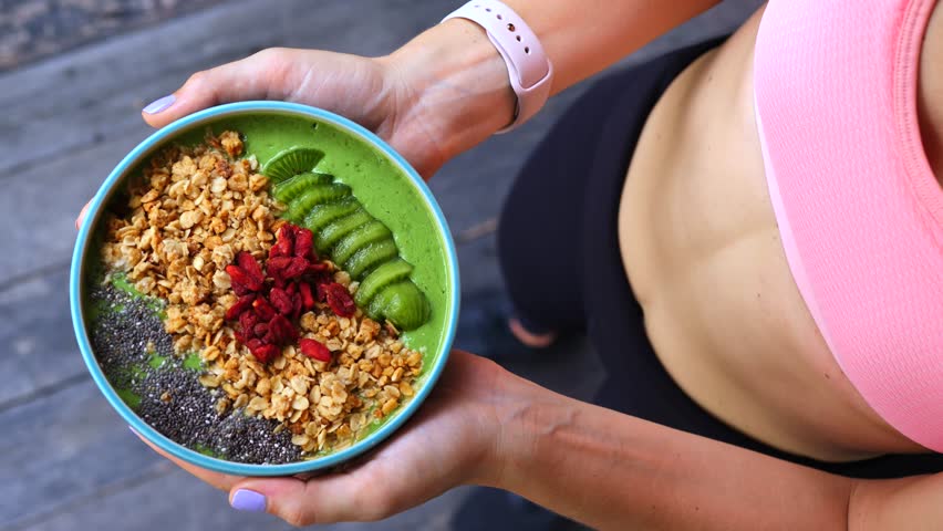 Top down view of a fit woman in sportswear holding a delicious green smoothie bowl topped with granola, chia seeds, kiwi, and goji berries, representing a healthy lifestyle and clean eating