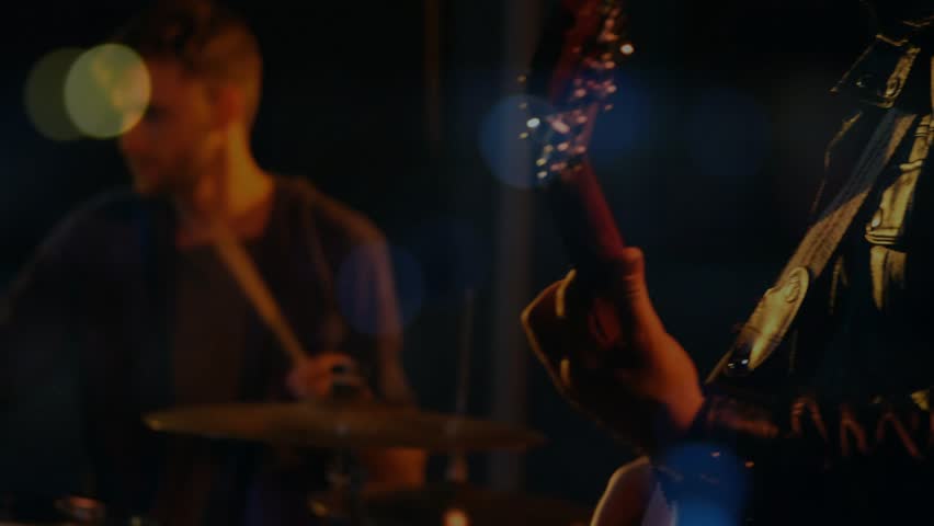 Drummer starting beat, camera cutting to lead singer singing into vintage mic, raising band energy. Backlit, bokeh, stage, moody, live, concert, ensemble