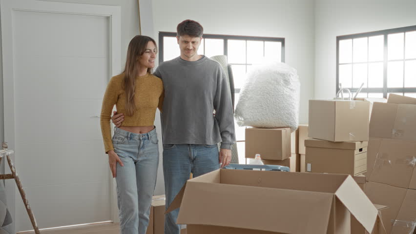Young couple embraces with arm around waist amid moving boxes in building, woman points ahead; hopeful togetherness.
