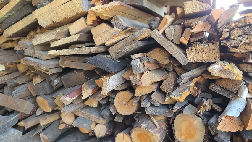 A large pile of chopped firewood and old wooden planks in a rural backyard. An axe is stuck in a wooden stump in the foreground