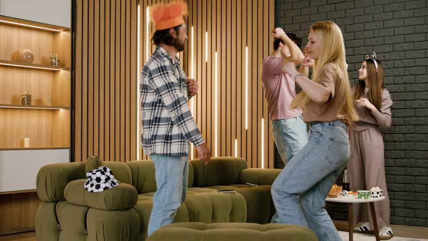 Group of friends celebrating in a modern living room, wearing festive hats and enjoying a lively atmosphere while dancing and playing with a soccer ball