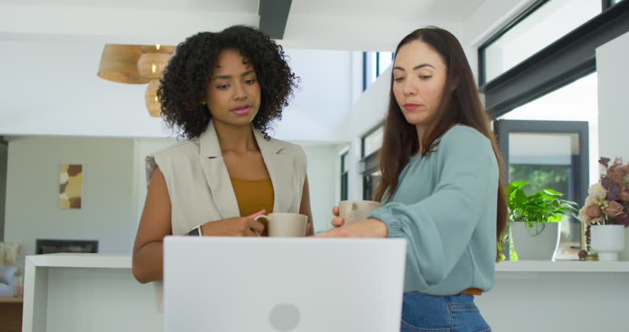 Diverse female coworkers standing at kitchen counter holding mugs pointing at laptop reviewing file. Collaboration, teamwork, computer, professional, modern, home, workspace