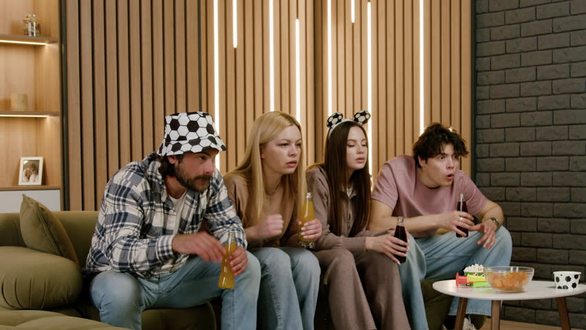 Group of friends celebrating a last-minute goal in a living room, raising drinks and cheering with excitement, wearing festive hats and surrounded by snacks