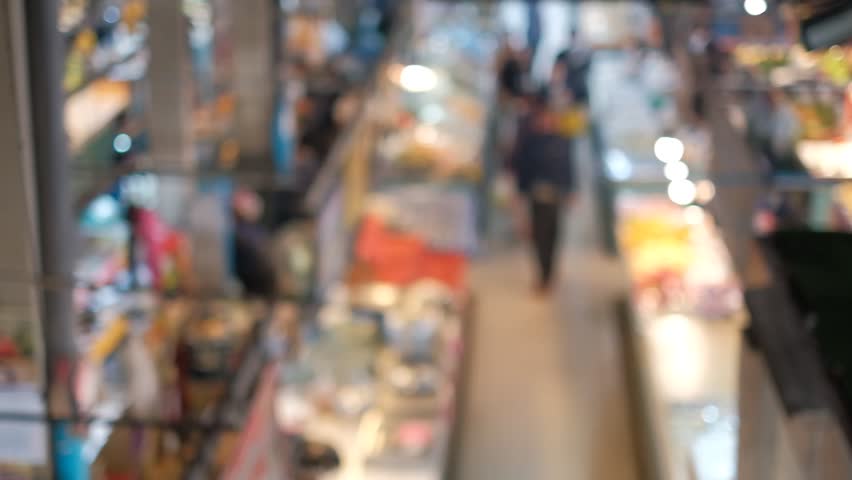 Blurred shoppers move through a lively market aisle. Shoppers stroll past colorful stalls and bright lights. Warm tones suggest a bustling, busy day