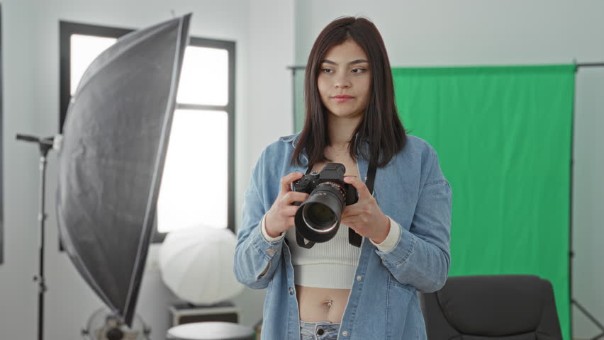 Woman holding dslr camera with strap, smiling and giving thumbs up in green screen photo studio; confidence warmth.