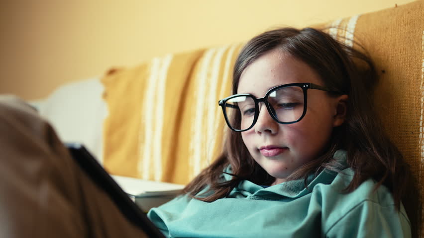 Little Girl in Glasses Using Digital Tablet on Couch with Stack of Books, Child Studying with Pad at Home, 4K 10-bit