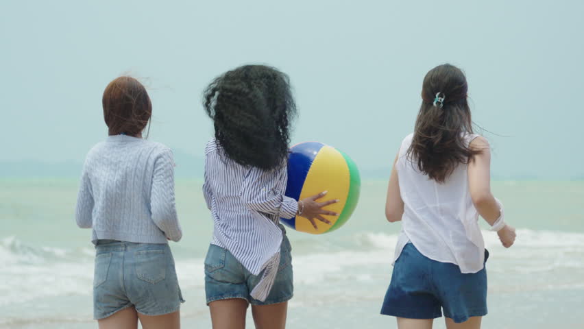 Joyful slow-motion video showing three diverse young women (Caucasian, Black, Asian) running and jumping together simultaneously on a sandy tropical beach during a sunny summer vacation.