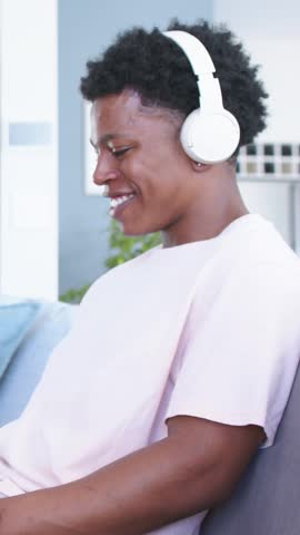 Vertical video: Hearing off-screen sound, man laughing on couch with white headphones, pink T-shirt. Sofa, plant, shelving, livingroom, home, stock, vertical video