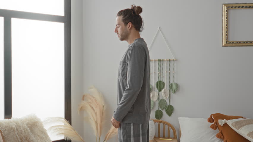 Man stretching arms beside bed in a sunlit bedroom in a residential building, reaching toward window while wearing pajamas and looking sideways; calm morning.