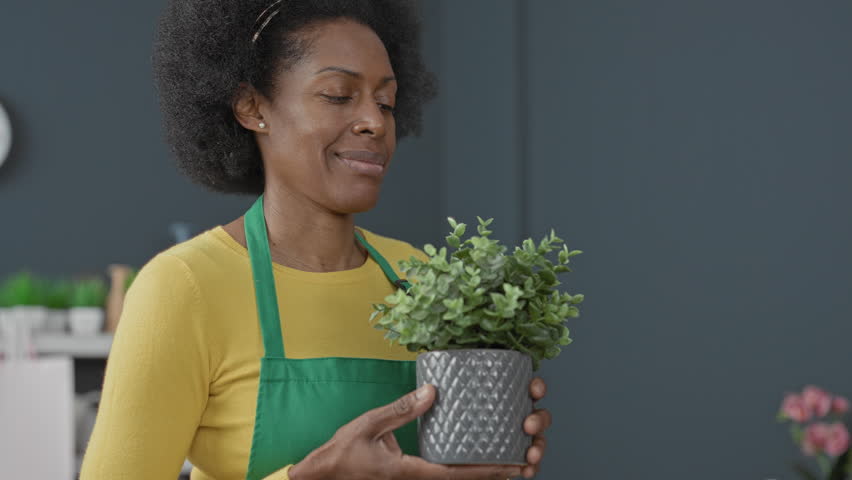 Woman florist wearing green apron holding a small potted plant with both hands in a bright flower studio workspace, smiling and presenting the pot; care pride joy.