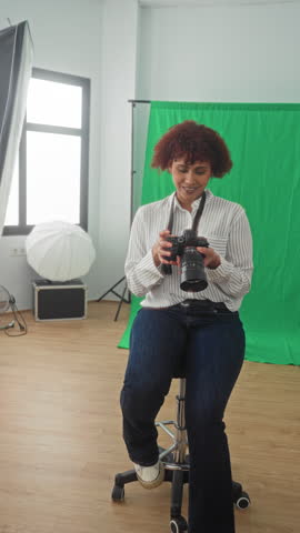 Woman photographer holding a camera with large lens, smiling and giving thumbs up in studio with green screen and softbox light; friendly confidence.