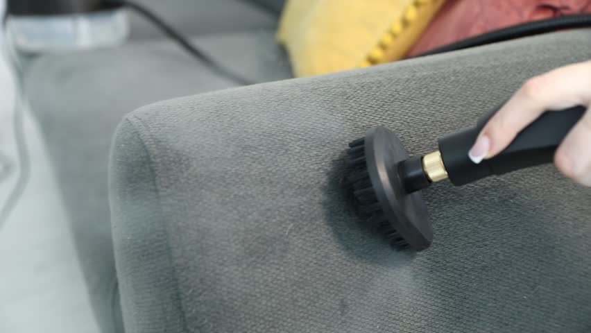 Woman cleaning grey sofa with steam cleaner at home, closeup. Camera moving right