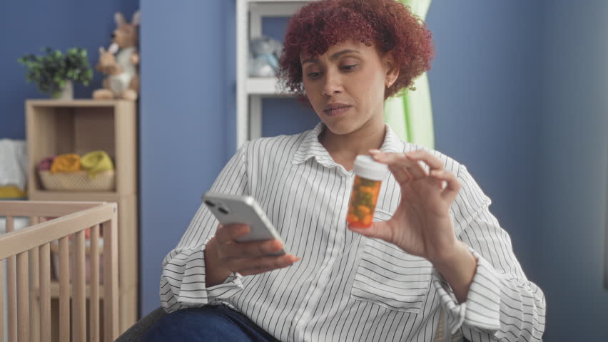 Woman holding smartphone and prescription bottle beside crib in nursery room checking label and reading dosage instructions; parental concern.