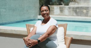 Senior woman sitting in wicker armchair at pool, adjusting sleeve and showing smartwatch to camera. Mature, outdoor, patio, sunlit, relaxed, chair, wearable - Powered by Shutterstock - Get 15% off with code: PIKWIZARD15