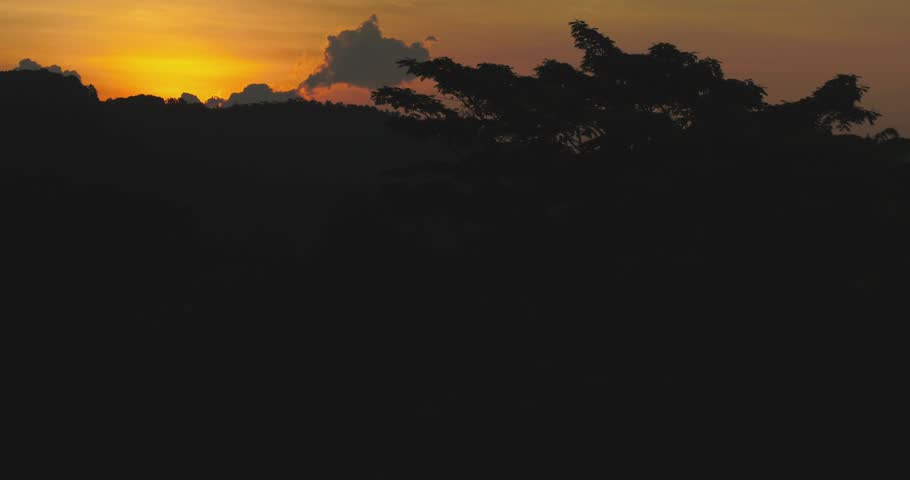 Cinematic aerial view of sunset over layered tropical mountains with sun rays piercing through the jungle canopy and misty atmosphere.