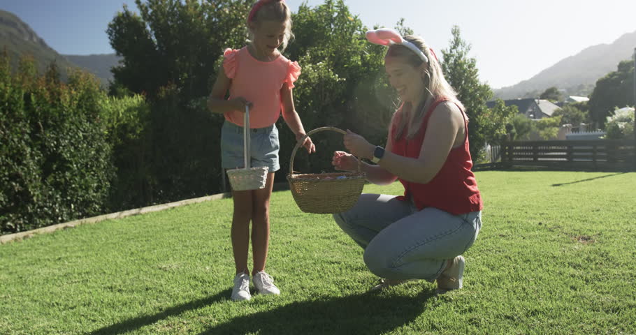 Mother reaching into wicker basket handing bright eggs to daughter filling white basket on lawn. Caregiver, child, backyard, grass, outdoor, sunny, residential