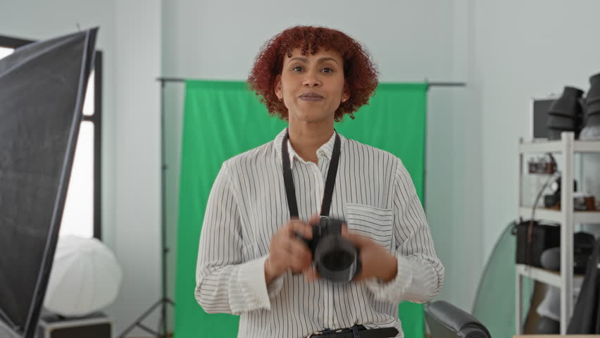 Woman holding camera and gesturing with hand in studio in front of green backdrop, wearing striped shirt and visible camera strap; confidence creativity.