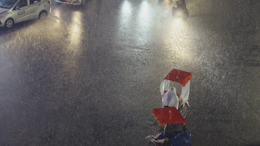 Two cyclos carrying passengers move through intense rain on busy Hanoi street with car headlights reflecting on soaked asphalt