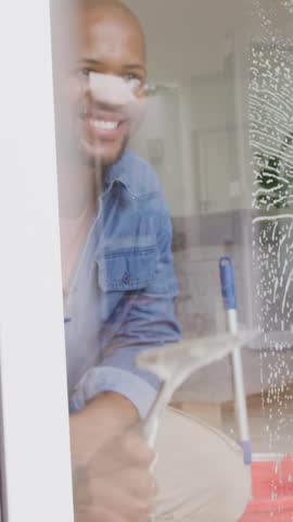 Vertical video: Preparing tools, man in denim shirt wiping home window with squeegee, removing suds. Glass, reflections, cleaning, interior, routine, sponge, vertical video