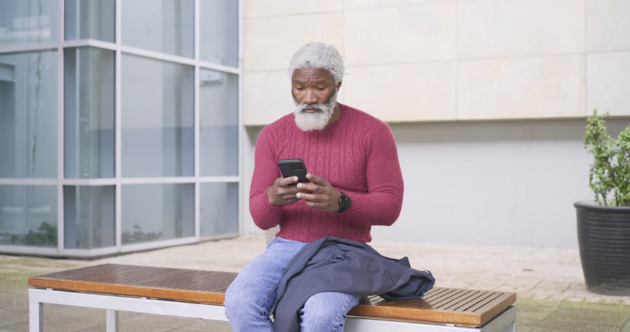 Senior African American man arriving, sitting on plaza bench, tapping phone, smiling, laughing. Urban, casual, contemporary, technology, leisure, expression, relaxed