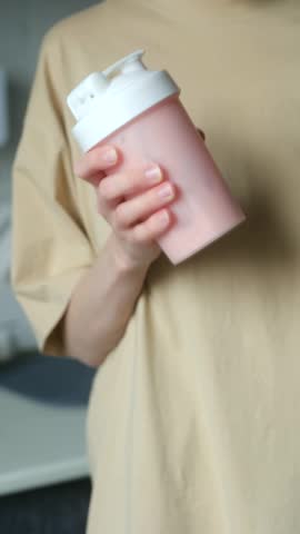 Unrecognizable woman in beige t shirt shaking pink bottle mix protein shake, diet supplement for fitness enthusiasts bodybuilders people leading healthy lifestyle. Woman making protein shake in bottle