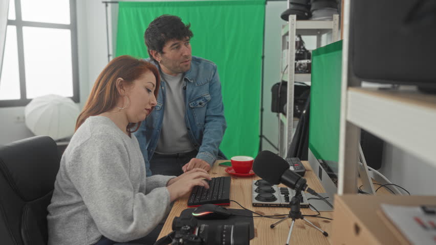 Man talking on phone hand to ear leans toward woman at green screen studio with keyboard and microphone; collaboration teamwork.