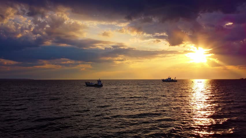 Cinematic view of a beautiful sunset over the Thermaikos Gulf in Greece with fishing boats. Dramatic sky with clouds, golden hour, and sun rays reflecting on the water.
