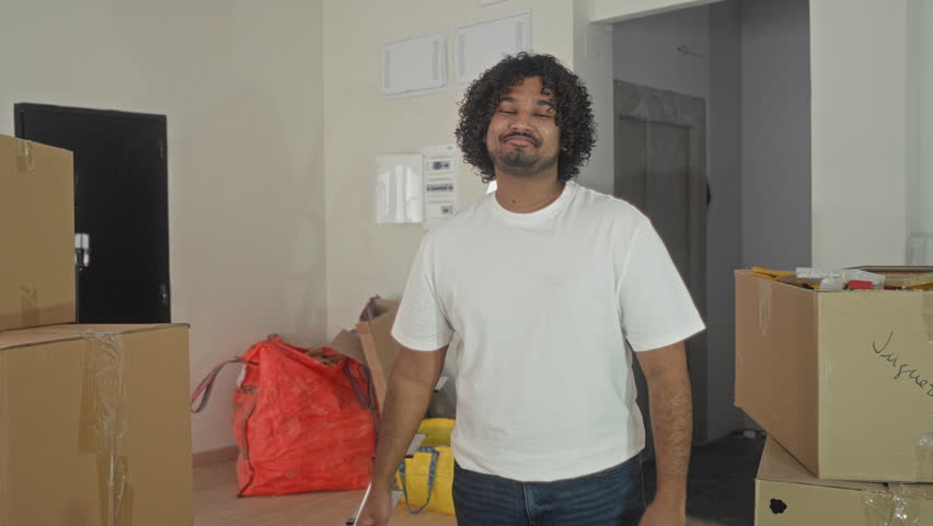 Man arms crossed smiling among cardboard boxes and a ladder in building new home interior; moving day pride.