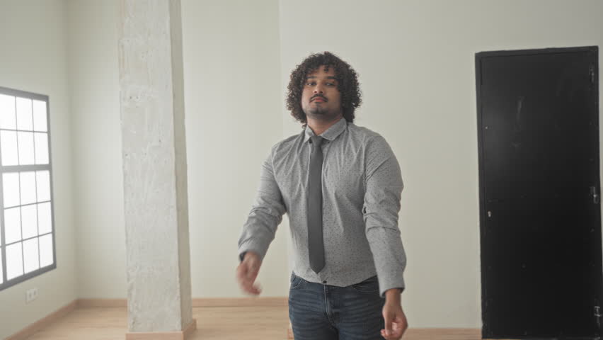 Man with arms crossed wearing a dotted shirt and tie, standing in an empty building room by a large window and concrete column; confidence trust.
