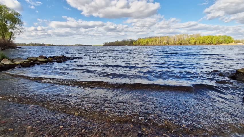 Picturesque riverbank with stones and blue sky with clouds. Rocky lakeshore with clear water under bright blue sky on a sunny day. Forest visible in the background. Natural landscape with bay