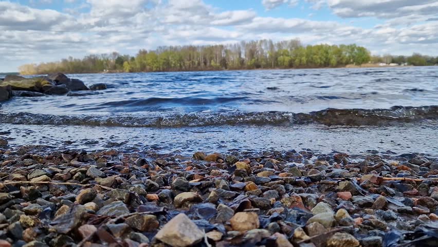 Picturesque riverbank with stones and blue sky with clouds. Rocky lakeshore with clear water under bright blue sky on a sunny day. Forest visible in the background. Natural landscape with bay