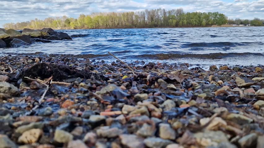 Picturesque riverbank with stones and blue sky with clouds. Rocky lakeshore with clear water under bright blue sky on a sunny day. Forest visible in the background. Natural landscape with bay
