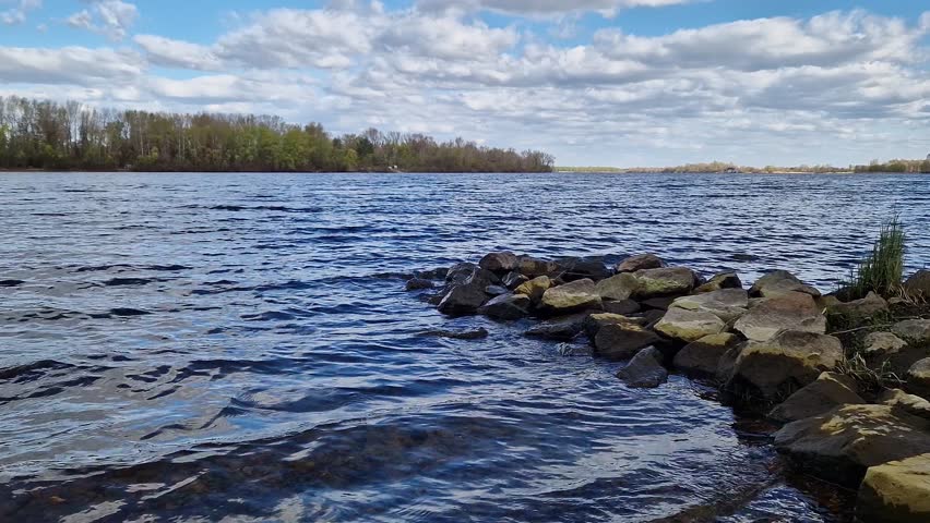 Picturesque riverbank with stones and blue sky with clouds. Rocky lakeshore with clear water under bright blue sky on a sunny day. Forest visible in the background. Natural landscape with bay