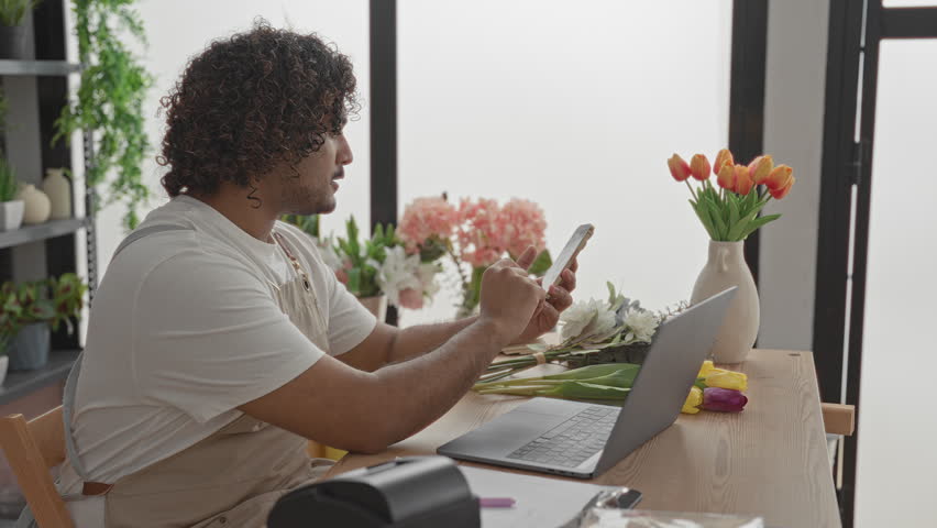 Man tapping smartphone and holding stems while arranging tulips at a shop table with laptop, vase and label printer in a studio; calm focus.