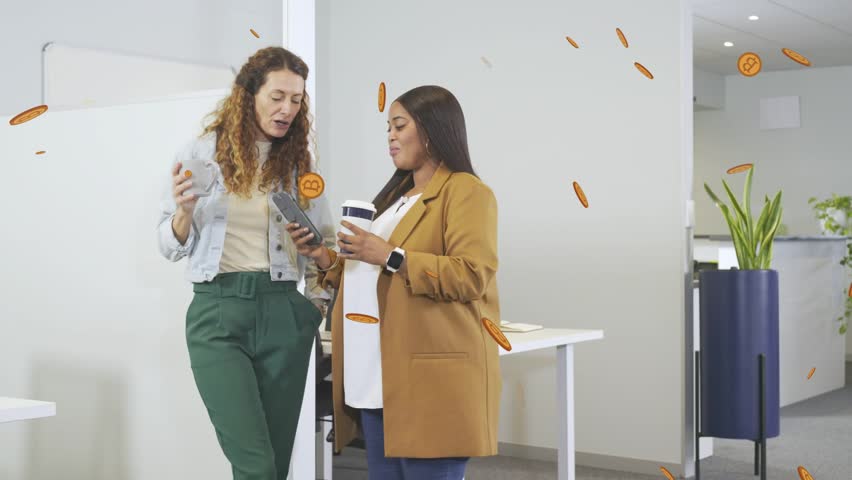 Right woman showing phone, sharing crypto, left woman leaning, sipping, bitcoin icons floating. Colleagues, office, collaboration, discussion, candid, modern, upbeat