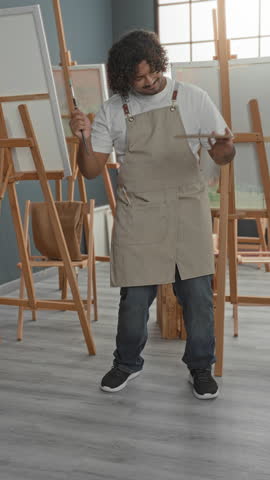 Man wearing apron raises brush above canvas while holding wooden palette and moving at easel in studio; creative joy.