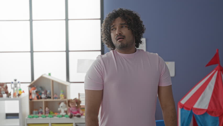 Man covering mouth while yawning, later with arms crossed, standing among toys, dollhouse and play tent in a bright kindergarten building; fatigue.